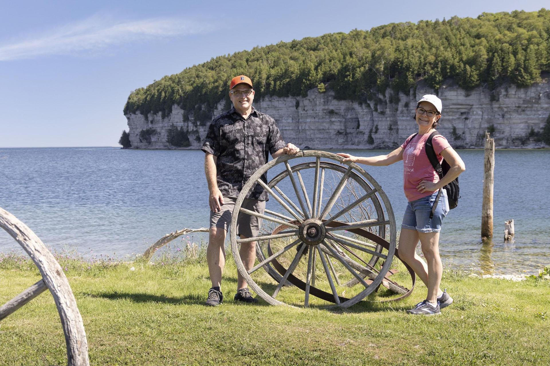 Two people pose for a photo beside an antique wheel, with the cliffs across Snail Shell Harbor in the distance.