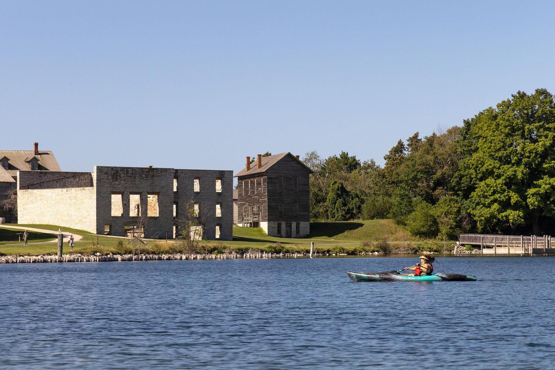 A kayaker paddles across Snail Shell Harbor, with historic buildings visible in the distance.